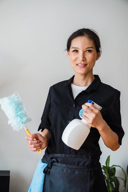 A professional cleaning expert from Carpet Cleaners W13 stands indoors in front of a plain light-colored wall, holding a spray bottle of cleaning solution in one hand and a cleaning brush in the other